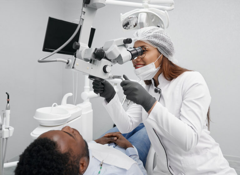 Positive female dentist in disposable hat, white uniform and gloves curing teeth of male patient. Clever doctor using professional equipment while african man lying on dentist chair.